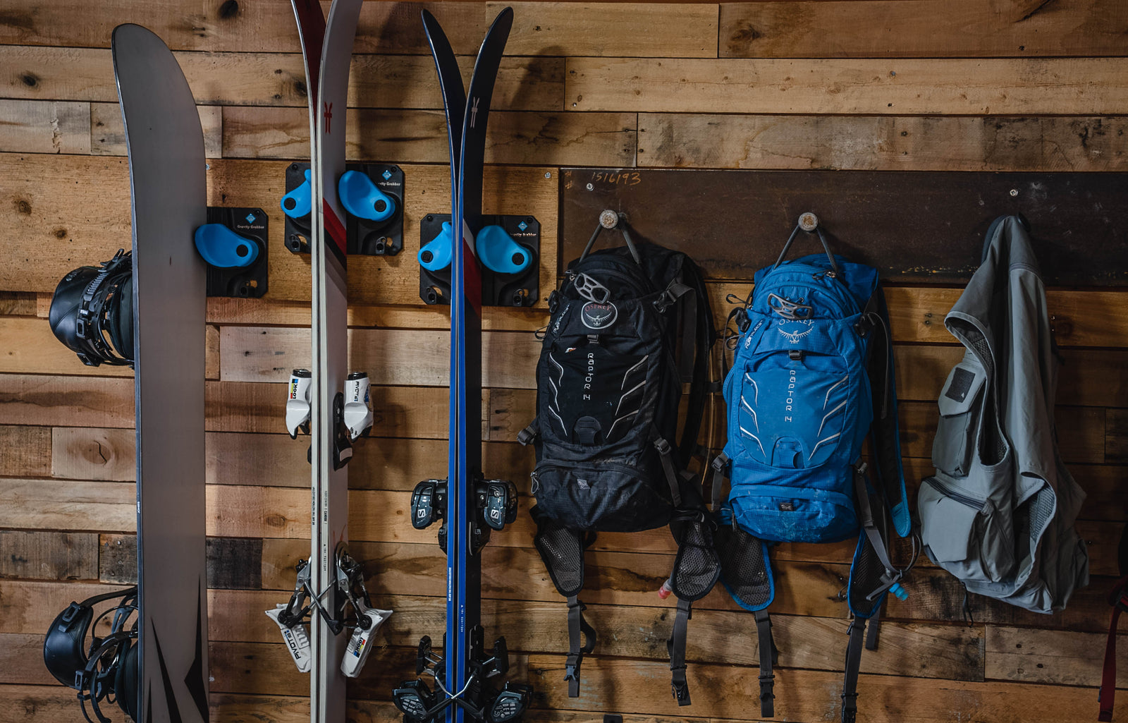 Wall-mounted Gravity Grabber racks holding skis and a snowboard next to backpacks and a jacket, neatly organized in a wooden garage wall setup.