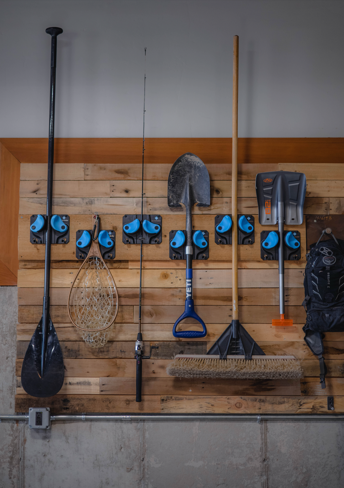Garage wall with multiple Gravity Grabber racks holding a paddle, fishing net, shovel, broom, and other outdoor tools neatly organized on a wooden panel.