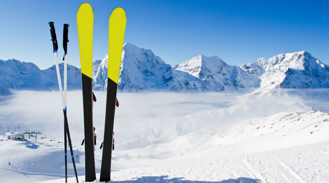A pair of skis and ski poles sit upright in the snow in front of snowy mountains.